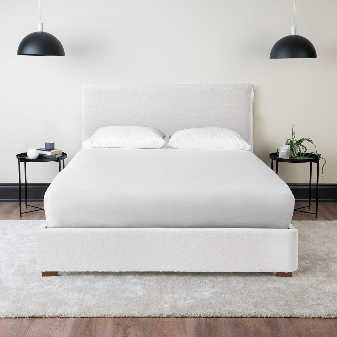 A white upholstered platform bed made up with white bedding in a bright bedroom with black pendant lights and bedside tables