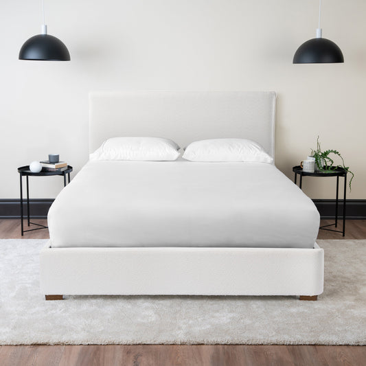 A white upholstered platform bed made up with white bedding in a bright bedroom with black pendant lights and bedside tables