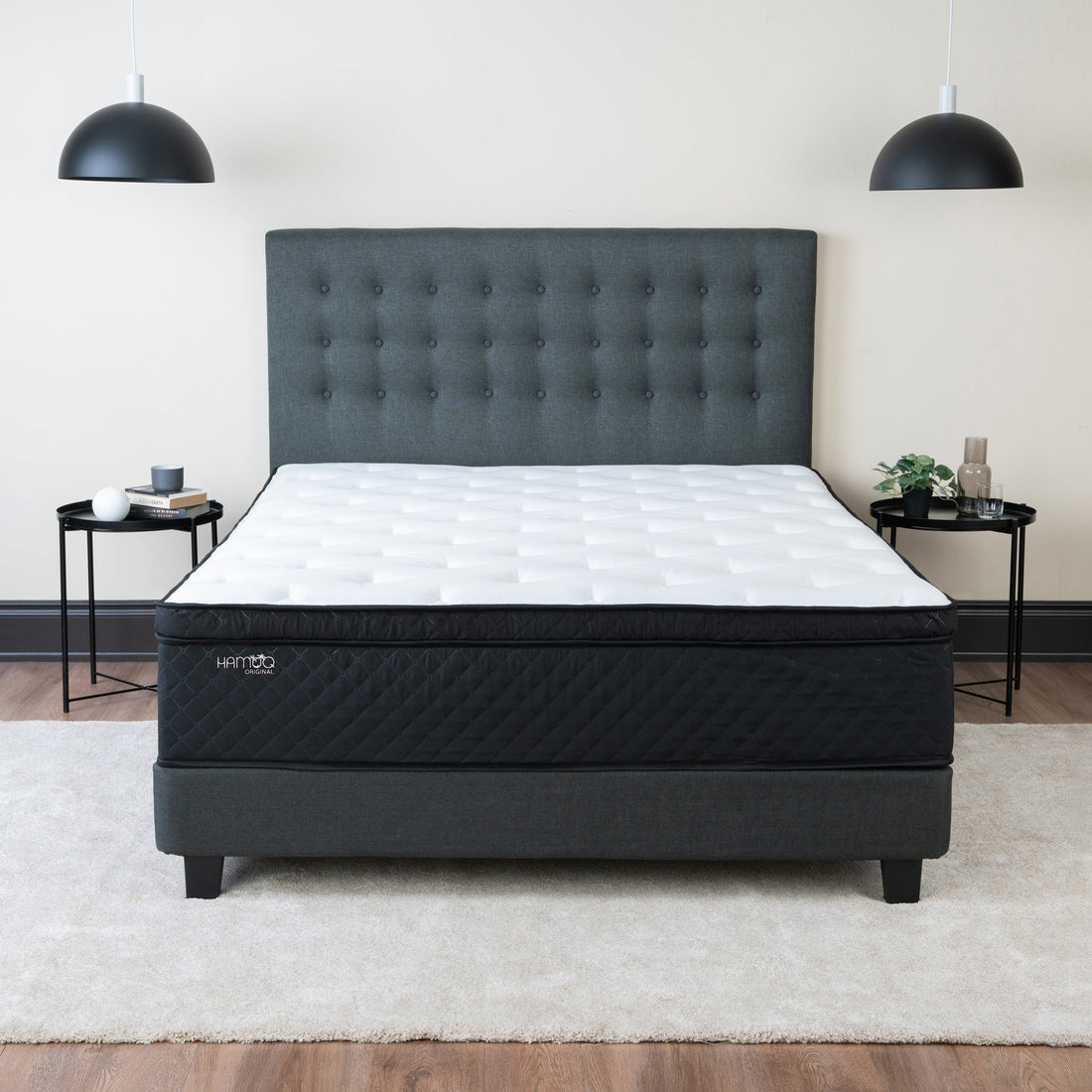 A white Hamuq hybrid mattress with a dark quilted border on a grey upholstered bed frame in a bedroom with two pendant lights
