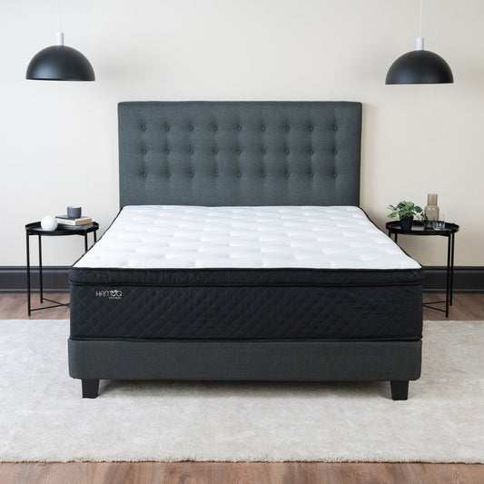 A white Hamuq hybrid mattress with a dark quilted border on a grey upholstered bed frame in a bedroom with two pendant lights